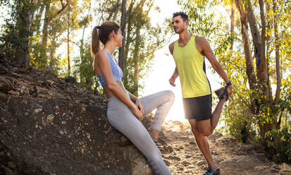 Fitte junge Frau in Sportkleidung sitzt auf einem Felsen und entspannt sich nach einem Trail-Run-Training mit einem Mann, der sein Bein ausstreckt. Mann und Frau, die eine Pause nach einem Lauftraining auf einem felsigen Bergpfad machen. - JLPSF18775