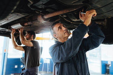 Zwei Mechaniker arbeiten unter einem Auto. Männer reparieren in einer Garage die Auspuffanlage eines gelifteten Autos. - JLPSF18785