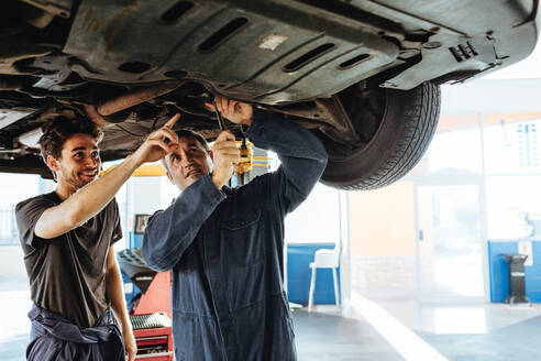 Mechaniker, der ein Auto repariert, mit einem Mitarbeiter, der auf ihn zeigt und lächelt. Zwei Autowerkstättenarbeiter arbeiten unter einem angehobenen Fahrzeug in einer Garage. - JLPSF18787