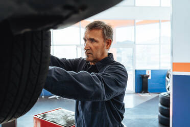 Mechanic fastening the wheel of a car in service station. Man replacing wheel of a vehicle lifted in garage. - JLPSF18789