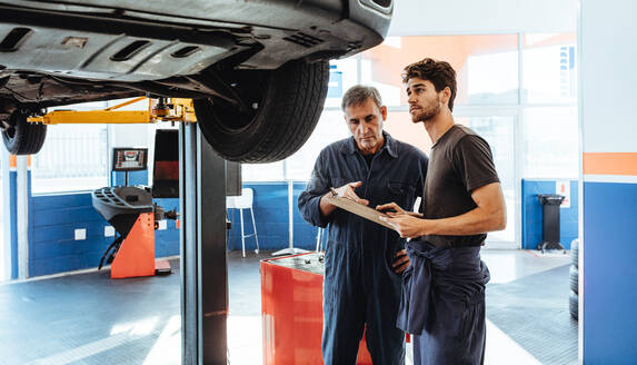 Zwei Automechaniker machen eine Liste auf einem Klemmbrett, während sie das Auto untersuchen. Mechaniker machen eine Liste von Reparaturen an einem Auto in einer Autowerkstatt. - JLPSF18793
