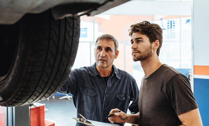 Two auto service professionals checking a car lifted on ramp and making notes. Mechanics inspecting the car in repair shop. - JLPSF18795