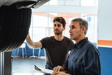 Mechanic showing the wheel of car to colleague making notes in clipboard. Auto service professionals examining the car and making notes. - JLPSF18796