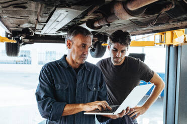 Automechaniker mit Laptop in der Autoreparaturwerkstatt mit einem Mitarbeiter, der daneben steht. Automechaniker mit Laptop bei der Untersuchung des Autos. - JLPSF18798