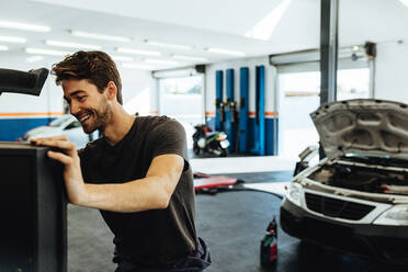 Lächelnder Mechaniker, der ein Auto mit einem Computer in einer Servicestation überprüft. Mechaniker, der eine computergestützte Diagnose eines Autos durchführt. - JLPSF18821