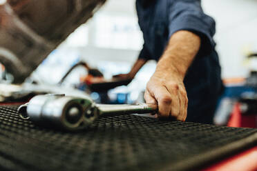 Hand eines Mechanikers, der einen Ratschenschlüssel vom Tisch in der Autowerkstatt aufhebt. Mechaniker arbeitet in der Garage mit Werkzeugen. - JLPSF18827