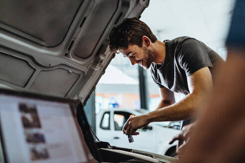 Lächelnder junger Mann bei der Autoreparatur in einer Garage mit einem Mitarbeiter, der einen Laptop benutzt. Ein Mechaniker arbeitet unter der Motorhaube eines Fahrzeugs. - JLPSF18829