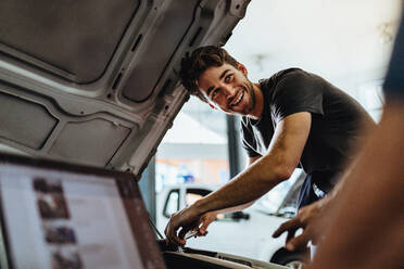 Automechaniker, der ein Fahrzeug in einer Tankstelle repariert. Automechaniker, der in einem Kfz-Servicezentrum arbeitet, schaut seinen Mitarbeiter an und lächelt. - JLPSF18830