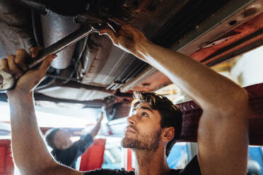 Young man working underneath a lifted car. Mechanic tightening a car part with spanner in automobile service center. - JLPSF18834