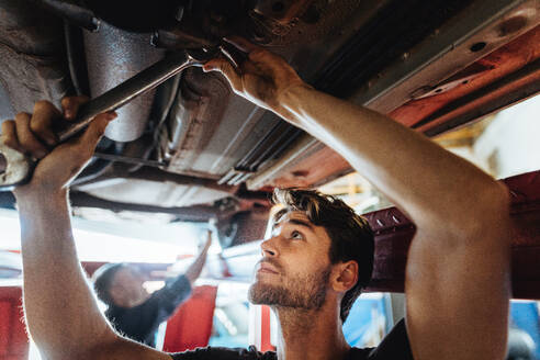Junger Mann bei der Arbeit unter einem angehobenen Auto. Mechaniker beim Festziehen eines Autoteils mit einem Schraubenschlüssel in einer Kfz-Werkstatt. - JLPSF18834