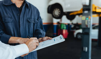 Ein Mechaniker nimmt das Dokument eines Kunden in der Werkstatt entgegen, der die Papiere unterschreibt, nachdem er sein Auto repariert hat. - JLPSF18840