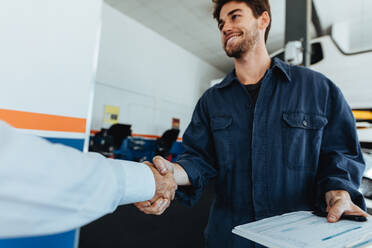 Young auto mechanic shaking hands with satisfied customer in garage. Automobile service center worker shaking hands with client after car servicing. - JLPSF18841