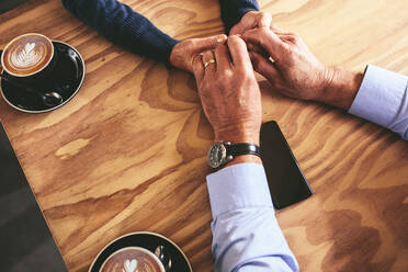 Close up of mature man and woman holding hands on a table with cups of coffee by the side. Top view of a senior couple sitting at a cafe. - JLPSF18852