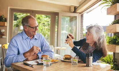 Smiling senior woman taking picture of her husband at a restaurant. Retired couple enjoying at a cafe clicking picutres with a mobile phone. - JLPSF18854