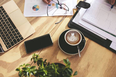 Draufsicht auf Tasse heißen Cappuccino Kunst Kaffee auf Holztisch mit Geschäftsleuten arbeiten auf einige Dokumente. Tasse Kaffee mit Milchschaum Kunst Muster auf Café Tisch mit Laptop, Handy und einige Papierkram auf Seite. - JLPSF18859