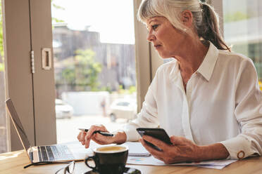 Senior businesswoman holding a smart phone sitting at coffee shop and working on laptop. Mature female entrepreneur at cafe using laptop computer. - JLPSF18860