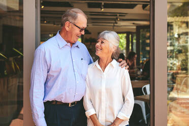 Smiling cafe owner couple standing at the door looking at each other and smiling. Senior man and woman at a restaurant. - JLPSF18862