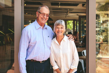 Successful cafe owner couple standing at the door with a digital tablet. Senior man wearing apron working at a restaurant. - JLPSF18863