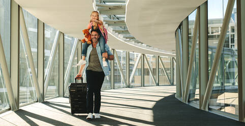 Mädchen sitzt auf der Schulter ihres Vaters am Flughafen. Vater und Tochter mit Gepäck gehen durch einen Tunnel, um ihren Flug zu besteigen. - JLPSF18872