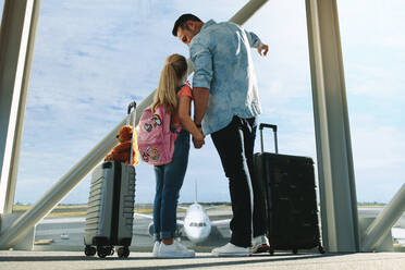 Rückansicht eines Vaters und seiner Tochter, die durch ein Fenster schauen, während sie auf ihren Flug am Flughafen warten. Ein Mann zeigt seiner Tochter ein Flugzeug, während sie am Flughafen-Terminal wartet. - JLPSF18874