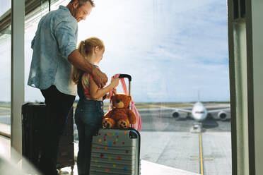 Vater und Tochter sehen sich Flugzeuge an, während sie im Flughafen warten. Mann mit seiner Tochter, die an einem großen Fenster im Flughafenterminal stehen. - JLPSF18875