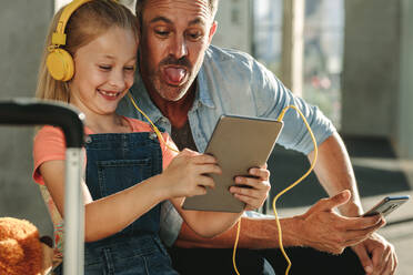 Niedliches Mädchen mit Kopfhörern, das auf ein digitales Tablet schaut, während ihr Vater die Zunge herausstreckt. Vater und Tochter machen lustige Videos auf einem digitalen Tablet, während sie in der Flughafen-Lounge warten. - JLPSF18878