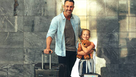 Vater und Tochter stehen zusammen mit ihrem Gepäck am Flughafen. Eine Familie, die zusammen reist, schaut in die Kamera und lächelt. Ein Mann mit einer Reisetasche und ein Mädchen mit einem Teddybär. - JLPSF18886