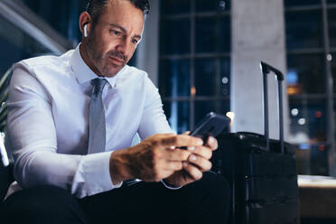 Businessman sitting at airport lounge and texting in his mobile phone. Business professional on a business trip using cell phone while waiting for his flight. - JLPSF18892