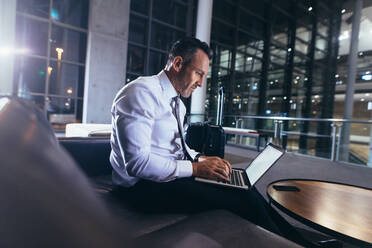 Geschäftsmann, der am Flughafen wartet und einen Laptop benutzt. Ein Mann in formeller Kleidung sitzt im Abflugbereich des Flughafens und benutzt seinen Laptop. - JLPSF18895