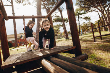 Zwei kleine Mädchen spielen auf einem Spielplatz im Freien. Zwillingsschwestern genießen es, auf einer Holzstruktur im Park zu spielen. - JLPSF18905