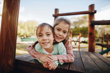 Kleines Mädchen umarmt ihre Zwillingsschwester, während in einer hölzernen Struktur auf dem Spielplatz liegen. Schöne kleine Mädchen genießen im Park spielen. - JLPSF18907