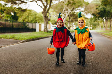 Zwei kleine Mädchen in Halloween-Kostüm Trick oder Behandlung auf der Straße. Zwei identische Zwillingsschwestern in Halloween-Kostüm zu Fuß im Freien mit Halloween-Eimer. - JLPSF18911