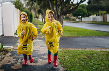 Zwei eineiige Zwillingsschwestern in gelben wasserdichten Mänteln und roten Gummistiefeln, die an einem regnerischen Tag draußen laufen. Kleine Mädchen, die bei Regenwetter draußen laufen. - JLPSF18927