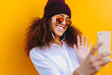 Urban african girl wearing hat and sunglasses doing a video chat using mobile phone. Attractive woman with curly hair waving at smartphone while having a video call against yellow background. - JLPSF18938