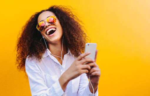 Lachende junge Frau, die über Kopfhörer Musik hört und dabei ein Handy benutzt. Afrikanisches Mädchen mit lockigem Haar und Sonnenbrille vor einer gelben Wand, das Lieder auf einem Smartphone hört. - JLPSF18944