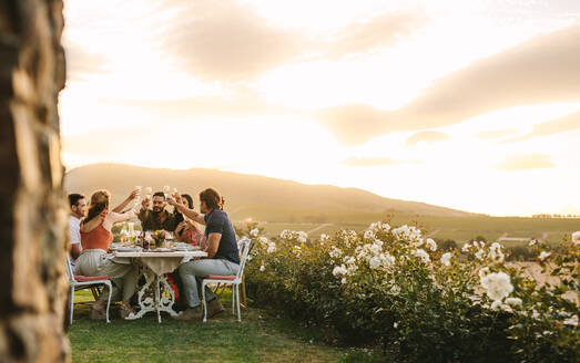 Eine Gruppe von Freunden stößt mit Champagnergläsern bei einer Dinnerparty im Freien an. Junge Leute trinken beim Abendessen im Garten. - JLPSF18945