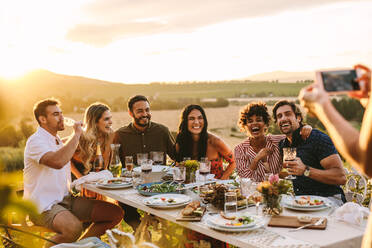 Eine Gruppe von Männern und Frauen posiert für ein Foto während einer Dinnerparty im Freien. Eine Frau fotografiert ihre Freunde bei einer Dinnerparty. - JLPSF18949