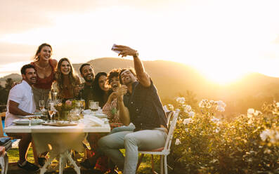 Freunde chillen draußen und machen ein Gruppen-Selfie. Lachende junge Leute sitzen um einen Esstisch im Freien und machen ein Selfie. - JLPSF18951