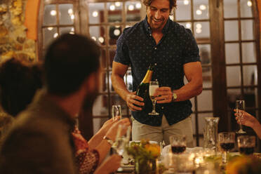 Mann serviert Freunden, die auf einer Party am Tisch sitzen, Getränke. Mann gießt bei einer Dinnerparty Champagner in eine Flöte. - JLPSF18969