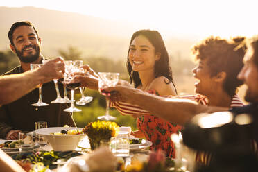 Eine Gruppe von Freunden stößt auf einer Party mit Getränken an. Junge Leute sitzen an einem Tisch, stoßen mit Getränken an und genießen das gemeinsame Abendessen. - JLPSF19001