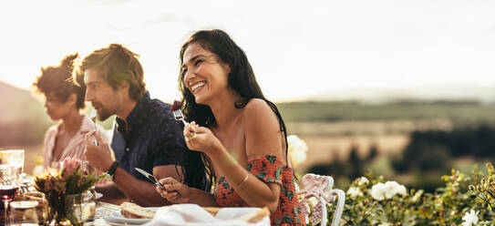 Lächelnde junge Frau beim Essen und im Gespräch mit Freunden während einer Dinnerparty im Freien. Frau genießt das Essen mit Freunden auf einer Party. - JLPSF19003