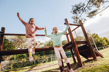 Happy little twin sisters jumping off a wooden log at the park Little girls having fun on outdoor playground. - JLPSF19015