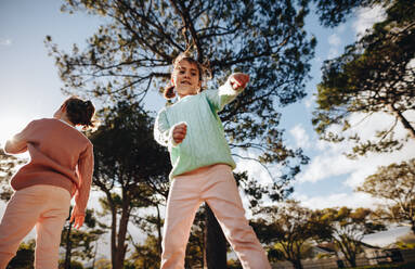 Niedriger Blickwinkel auf ein kleines Mädchen, das im Park tanzt, während ihre Schwester daneben steht. Kleine Mädchen haben Spaß auf dem Spielplatz im Freien. - JLPSF19017