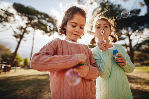 Hübsche kleine Mädchen spielen mit Seifenblasen auf dem Spielplatz. Süße Zwillingsschwestern blasen Seifenblasen im Freien. - JLPSF19018