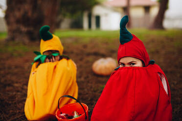 Two cute little girls sitting on ground hiding a in pumpkin costume with halloween bucket. Twin sisters tricking outdoors on halloween - JLPSF19026