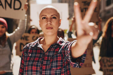 Glatzköpfige Frau protestiert im Freien und zeigt ein Friedenshandzeichen. Frau mit einer Gruppe von Menschen, die im Freien auf einer Straße protestieren. - JLPSF19036