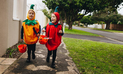 Identical twin sisters in halloween costume with halloween bucket walking on sidewalk. Halloween kids trick or treating. - JLPSF19040