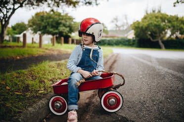 Beautiful young girl wearing helmet sitting in a red wagon cart by the road outdoors. Cute little girl in a toy trolley outdoors. - JLPSF19056