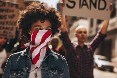 Porträt einer schweigend protestierenden jungen Frau mit einer Gruppe im Hintergrund. Afrikanische Frau, die ihren Mund mit einem Schal bedeckt und auf der Straße mit einer Gruppe weiblicher Aktivisten protestiert. - JLPSF19057