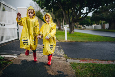 Zwei fröhliche Mädchen in gelben Regenmänteln und roten Gummistiefeln, Zwillingsschwestern, die in wasserdichten Mänteln im Freien spazieren gehen. - JLPSF19074